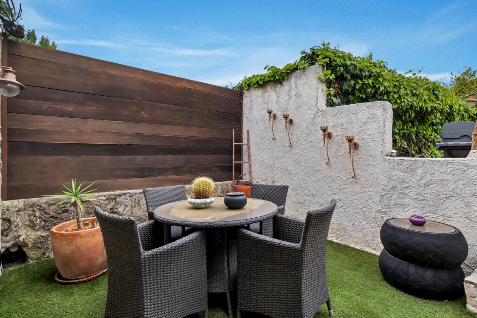 Private Terrasse mit Kunstrasen, runder Sitzgruppe aus dunklem Polyrattan und Wänden aus strukturiertem Putz, Holzpaneelen und Naturstein.