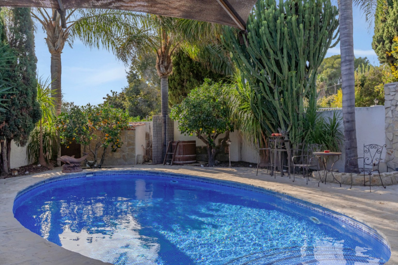 Privater Pool mit blauen Mosaikfliesen, umgeben von einem mediterranen Garten mit Palmen, Zitrusbäumen und einer Sitzecke aus Schmiedeeisen auf Naturstein.