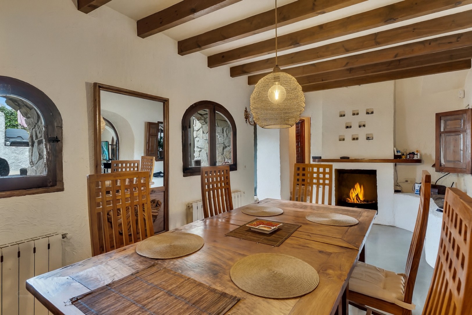 Rustikaler Essbereich mit Sichtbalkendecke, massivem Holztisch und offenem Kamin. Rundbogenfenster und eine geflochtene Pendelleuchte unterstreichen das Ambiente.