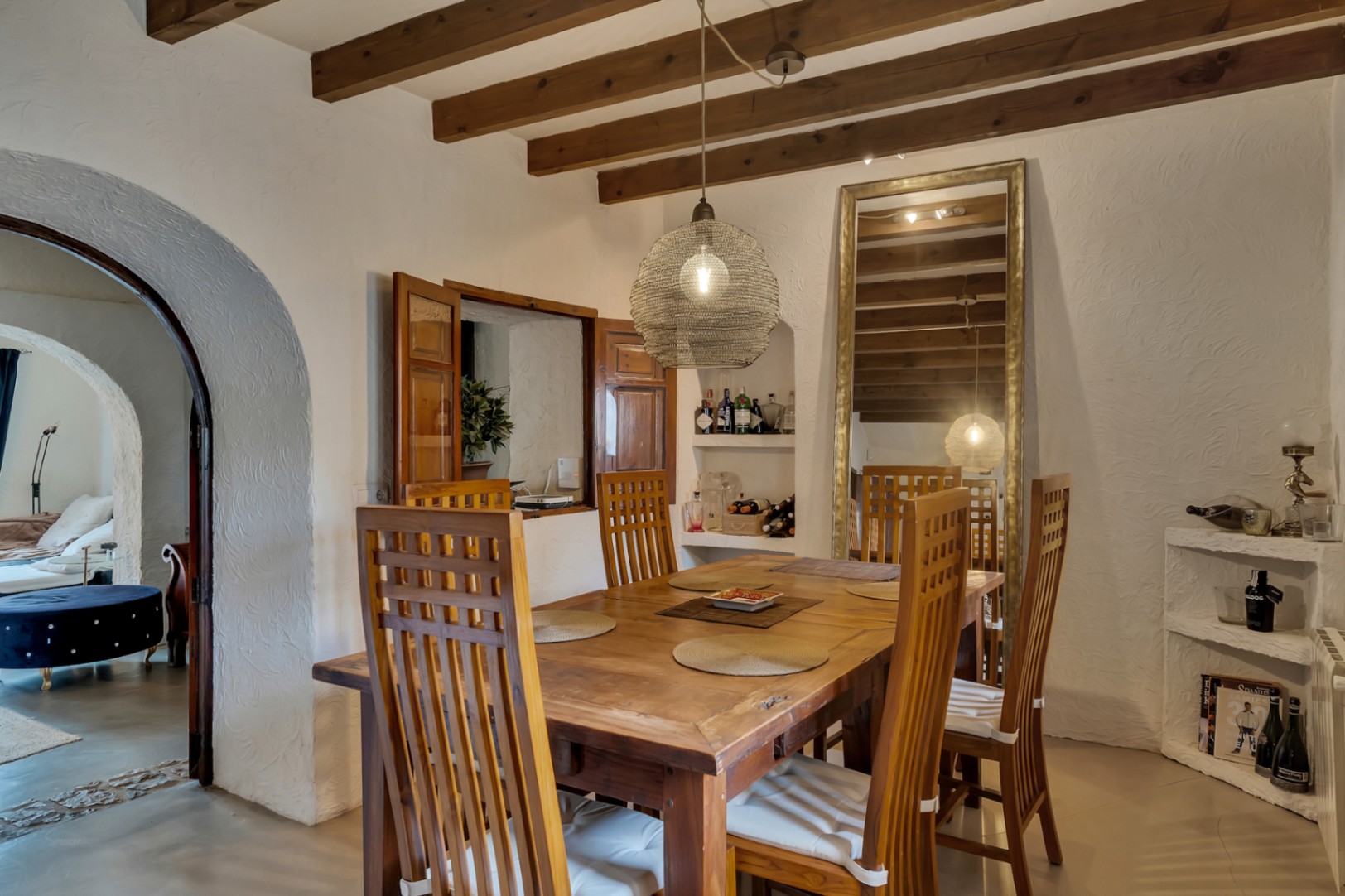 Rustikaler Essbereich mit sichtbaren Holzbalken, Massivholztisch und hohen Stühlen. Mit Durchreiche mit Holzläden und einem Rundbogen, der in den Wohnbereich führt.