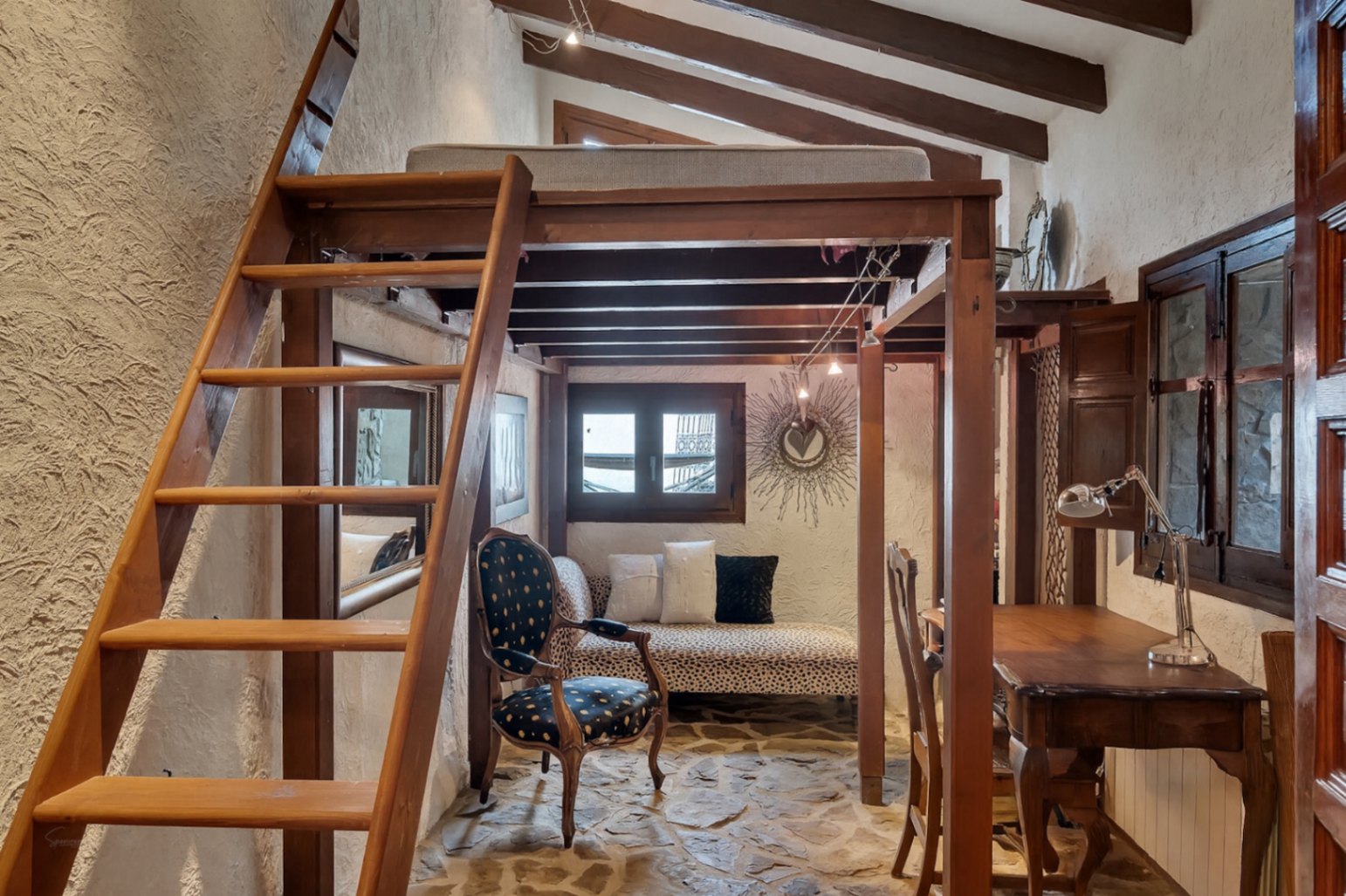 Rustikales Schlafzimmer mit hölzerner Hochebene, sichtbaren Deckenbalken und Natursteinboden. Unter der Empore befinden sich ein Sitzbereich und ein Schreibtisch.