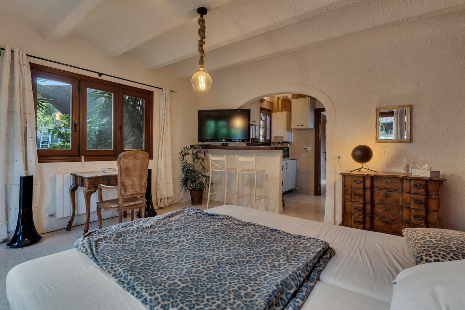 Schlafzimmer mit hölzernem Arbeitsbereich, Sichtbalkendecke und Rundbogen-Durchgang zu einer Pantryküche mit Frühstückstheke.
