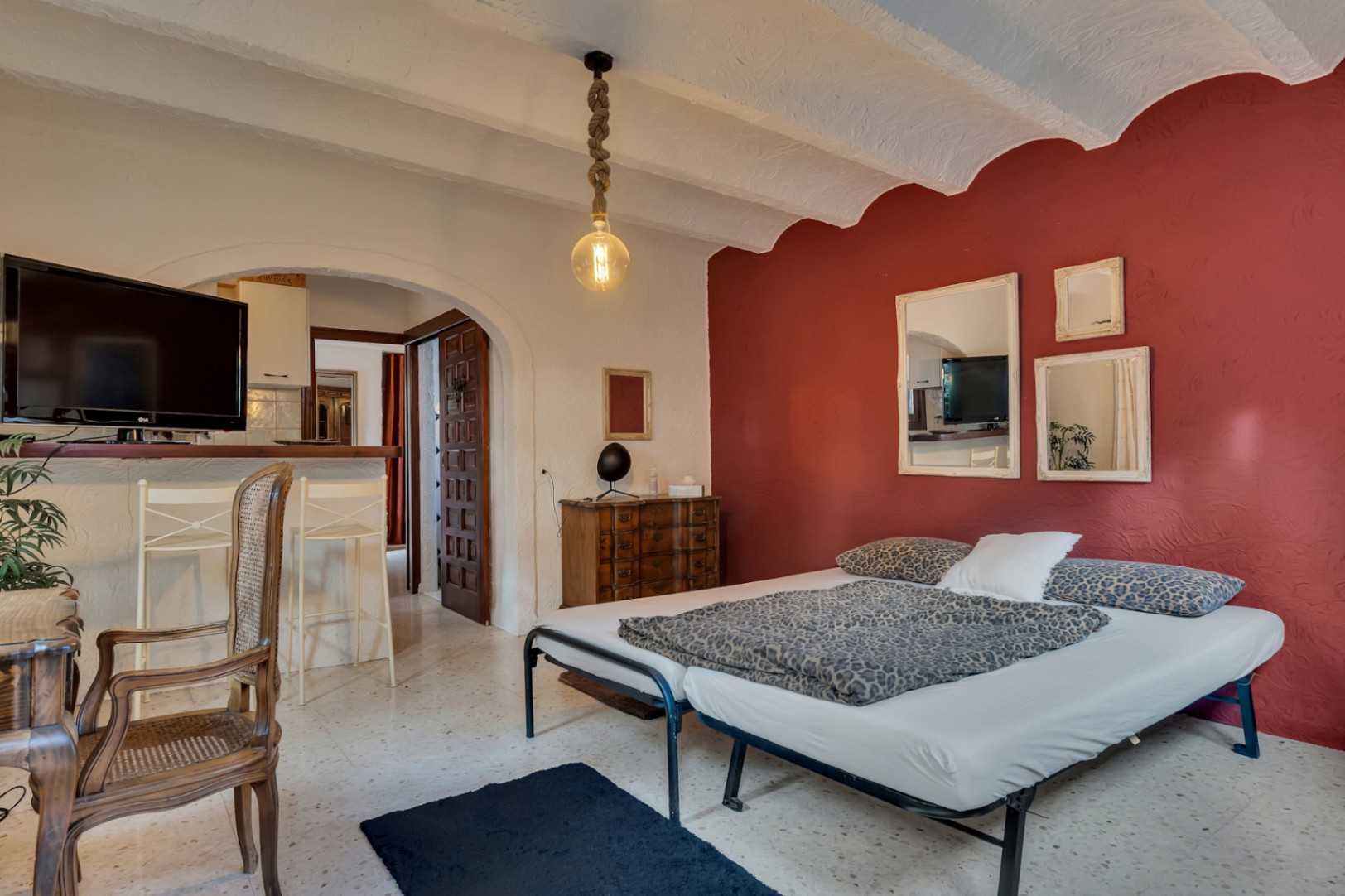 Schlafzimmer mit roter Akzentwand, gewölbten Decken und Terrazzoboden. Ein Rundbogen führt zum Wohnbereich, ergänzt durch eine rustikale Seilpendelleuchte.