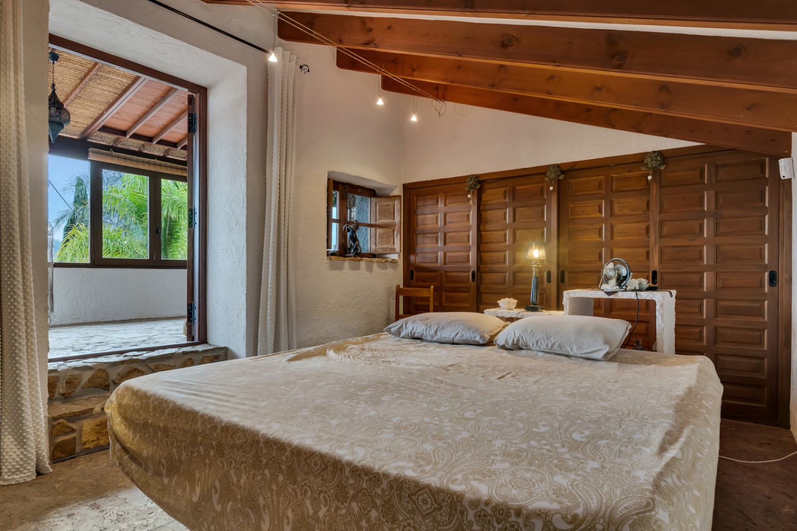 Schlafzimmer mit Sichtbalkendecke aus Holz, traditionellen Einbauschränken und direktem Zugang zu einer überdachten Terrasse über eine Steinstufe.
