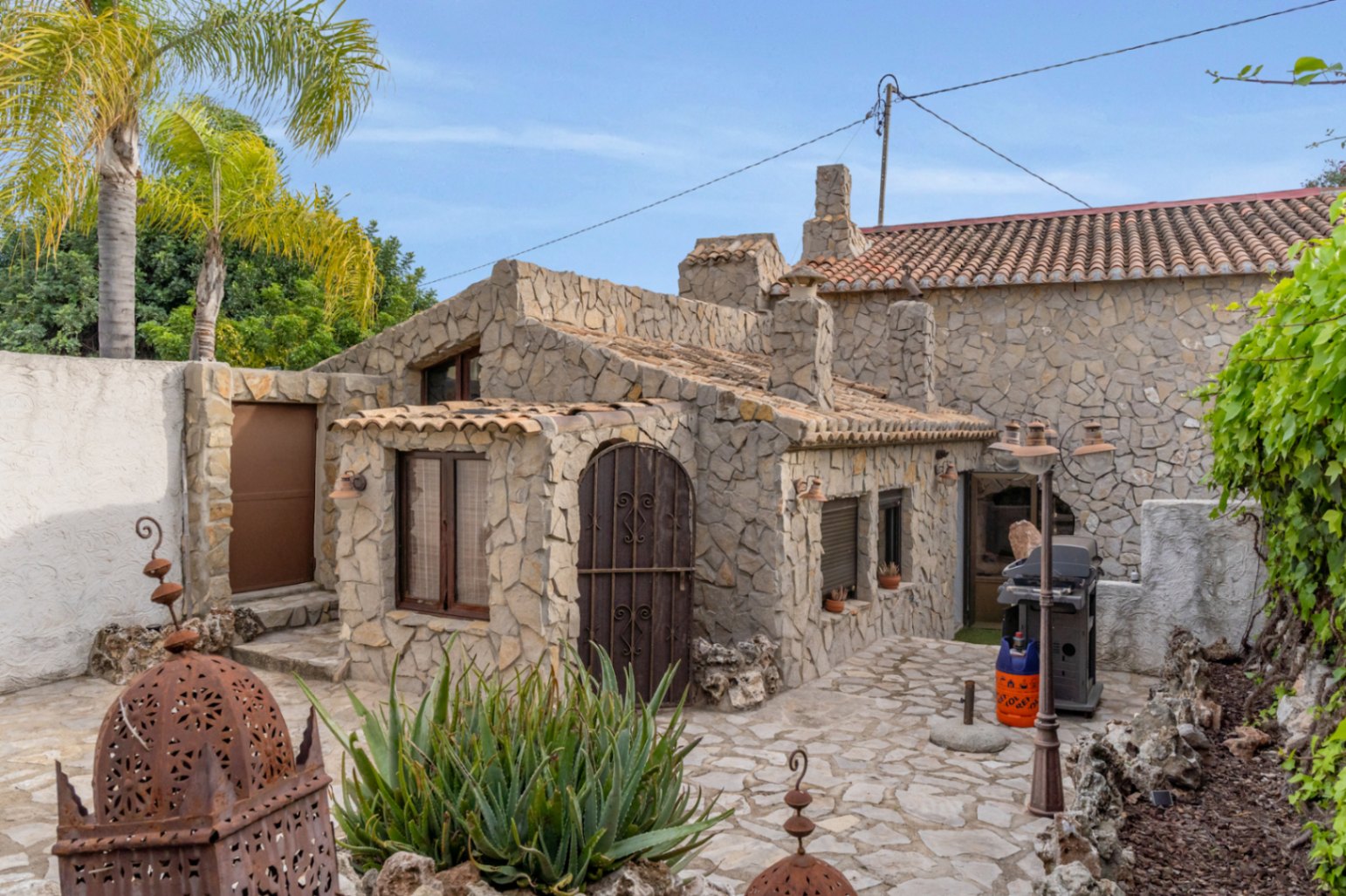 Separates Gästehaus mit Natursteinfassade, Terrakottadach und gepflasterter Terrasse inklusive Grillplatz und mediterraner Bepflanzung.