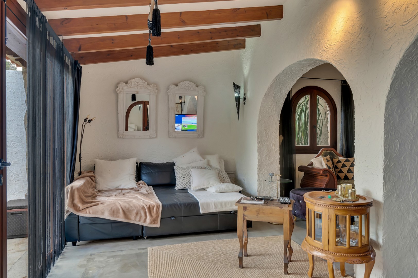 Wohnbereich mit sichtbaren Holzbalkendecken, strukturierten weißen Wänden und Rundbögen. Ausgestattet mit Ledersofa, verzierten Spiegeln und rustikalen Holzmöbeln.