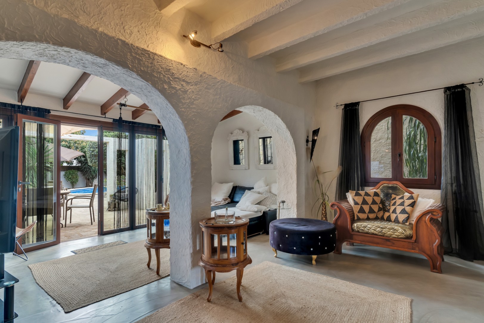 Wohnbereich mit traditionellen weißen Rundbögen, Holzbalkendecken und geschnitzten Möbeln. Große Glastüren führen direkt zur Terrasse und zum privaten Pool.
