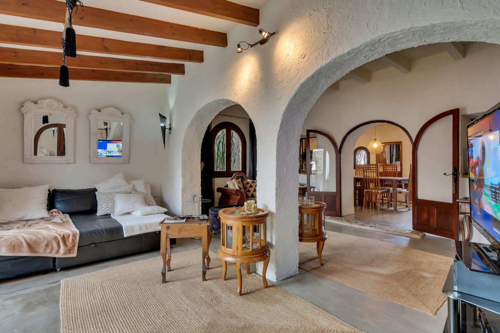 Wohnzimmer mit sichtbaren Deckenbalken aus Holz, traditionellen Bögen und strukturierten weißen Wänden, die durch verglaste Bogentüren zum Essbereich führen.