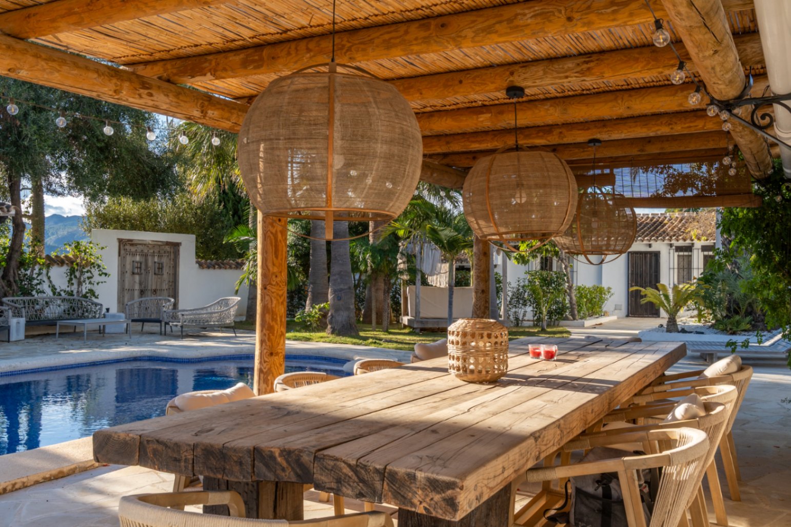 Außen-Essbereich unter einer Pergola aus Holzbalken und Schilfrohr, mit rustikalem Tisch, Naturfaser-Hängeleuchten und Blick auf den Pool und den mediterranen Garten.