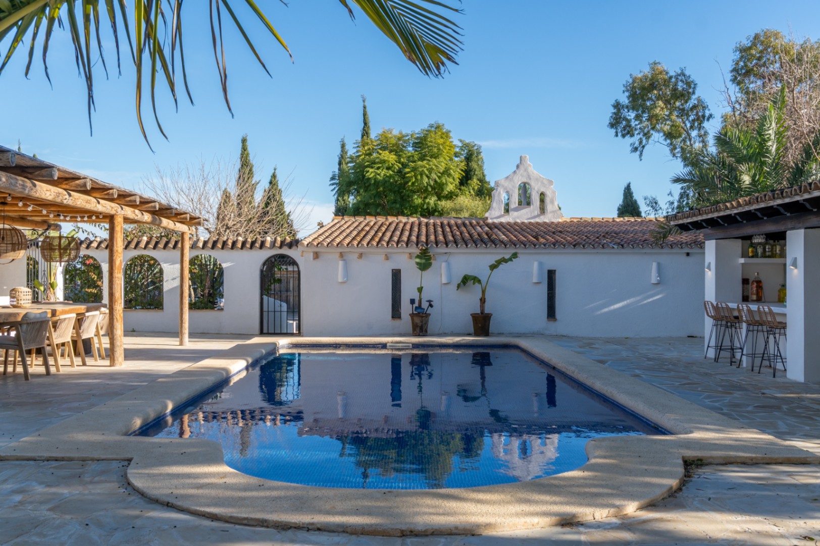 Außenbereich mit Pool, Steinterrasse, Holzpergola und einer Bar, ergänzt durch traditionelle mediterrane Mauern und eine Glockengiebel-Struktur.