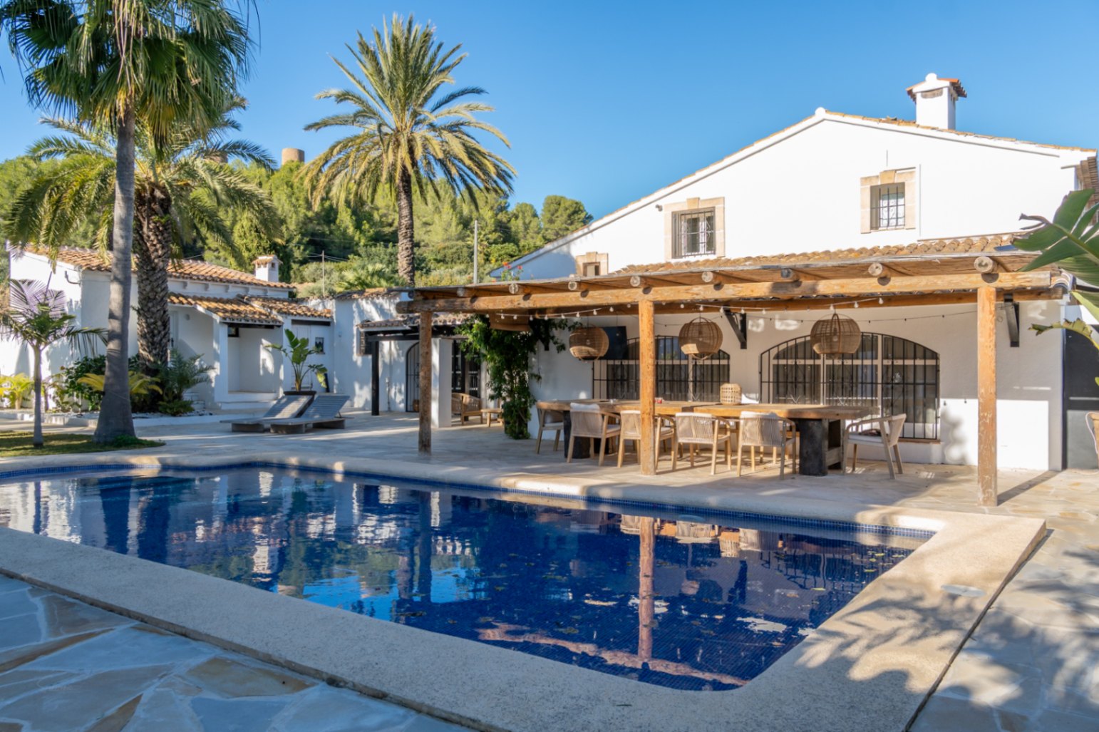 Außenbereich mit rechteckigem Pool und Holzpergola. Der Essbereich im Freien bietet einen massiven Holztisch, Korblampen und Blick auf die weiß getünchte mediterrane Finca.