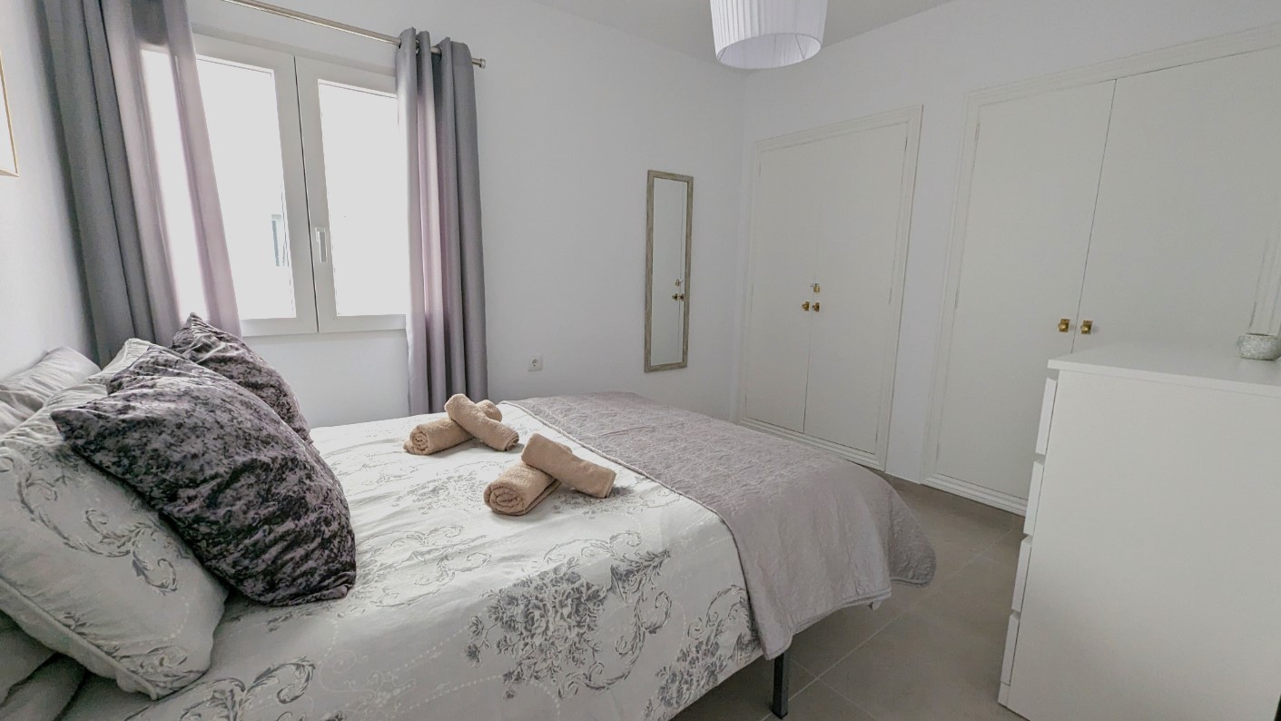 Doppelzimmer mit weißen Einbauschränken, Wandspiegel und Fliesenboden. Der Raum bietet natürliches Licht durch das Fenster und eine Einrichtung in neutralen Grautönen.
