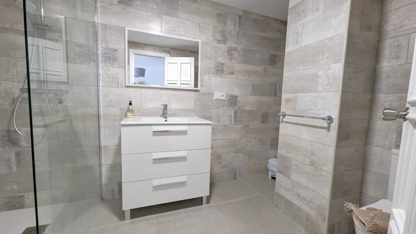Modernes Badezimmer mit weißem Waschtisch mit drei Schubladen, begehbarer Dusche mit Glastrennwand und strukturierten Wandfliesen in neutralen Beigetönen.