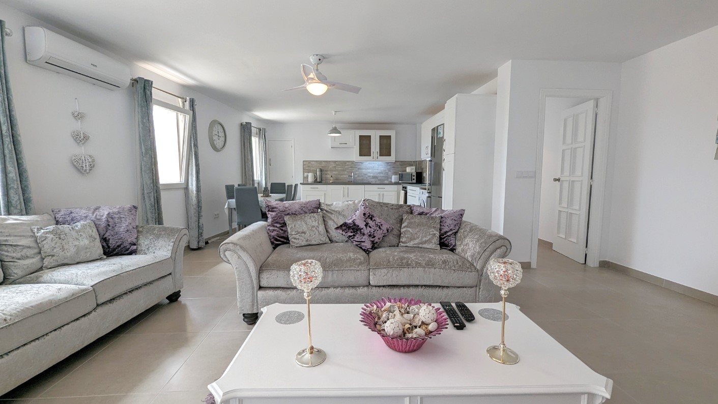Offener Wohnbereich mit grauen Samtsofas, weißem Couchtisch und Deckenventilator. Der Raum umfasst einen Essbereich und eine integrierte weiße Küche mit Fliesenboden.