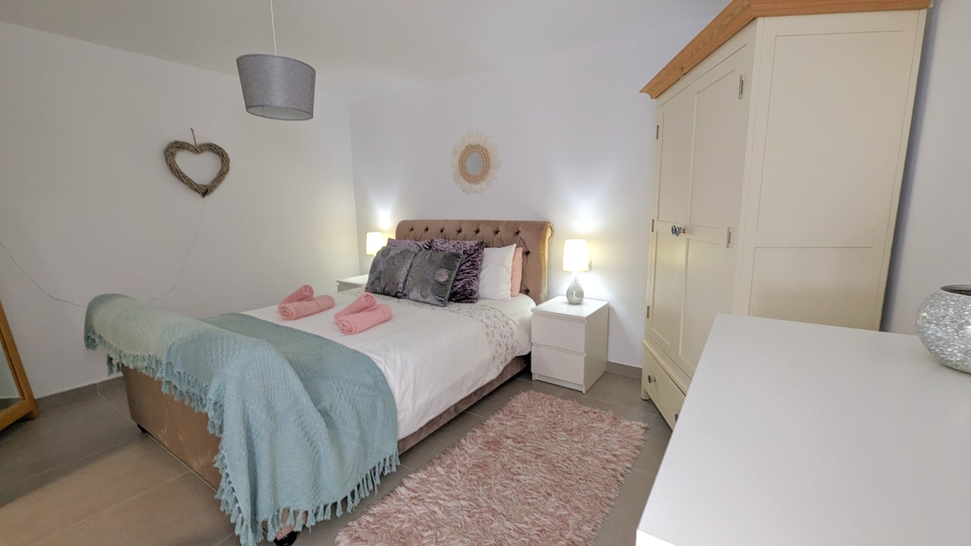 Schlafzimmer mit Doppelbett, gepolstertem Kopfteil und weißen Nachttischen. Ausgestattet mit einem cremefarbenen Kleiderschrank, Fliesenboden und dezenter Dekoration.