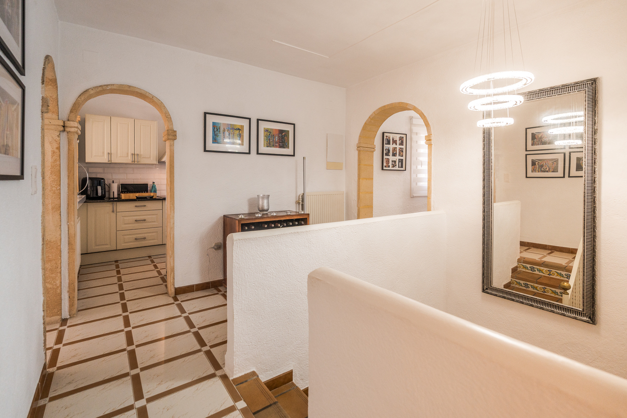 Flur mit Tosca-Steinbögen, Fliesenboden und modernem Kronleuchter. Sichtbar sind der Küchenzugang und eine Treppe mit traditionell dekorierten Setzstufen.
