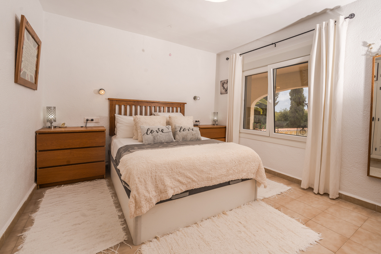 Helles Schlafzimmer mit Holz-Kopfteil, doppelt verglasten Fenstern und Fliesenboden. Ausgestattet mit passenden Holzkommoden und Blick auf den Garten.