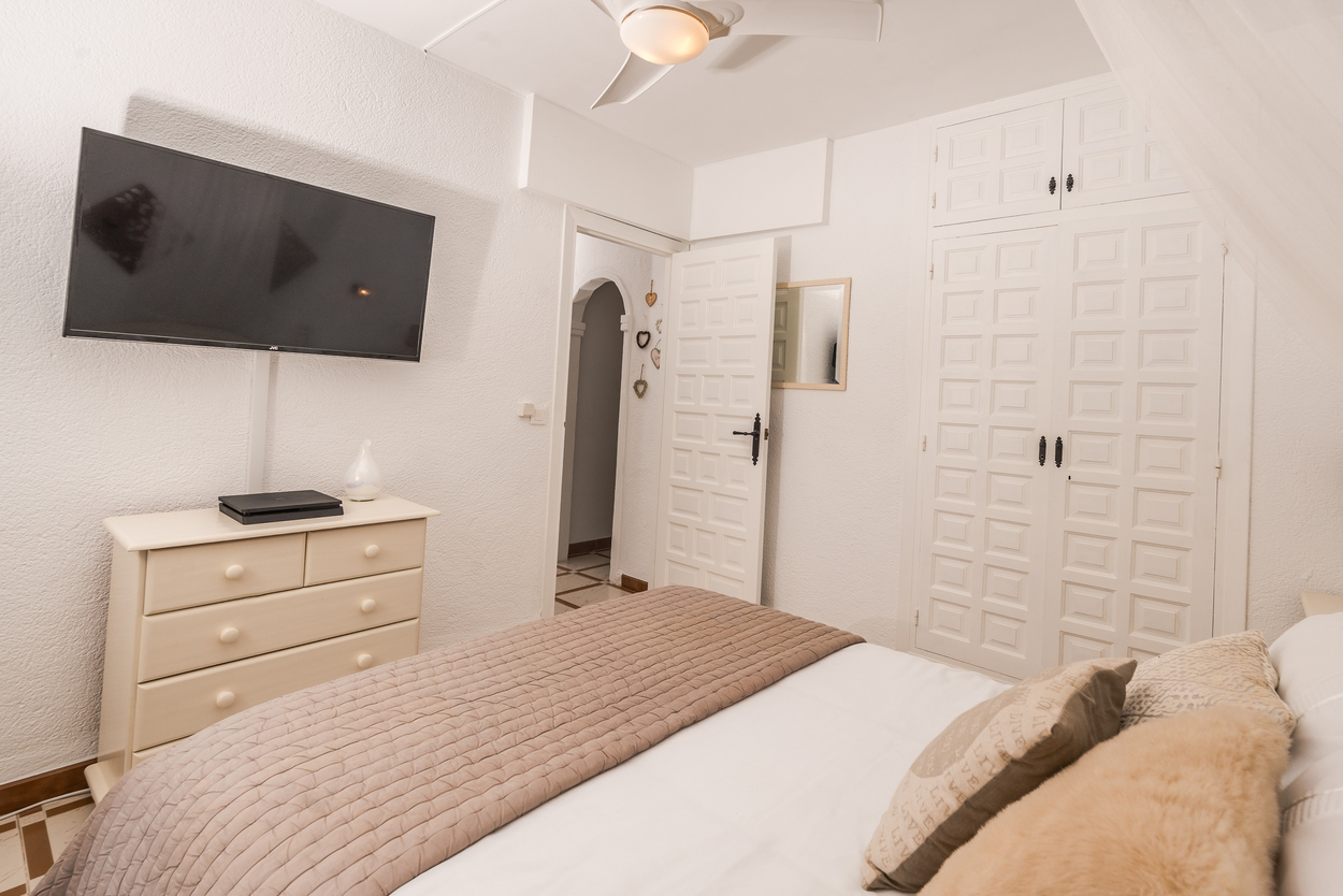 Helles Schlafzimmer mit weißen Einbauschränken, Wandfernseher und Deckenventilator. Ausgestattet mit passender Kommode und Fliesenboden.
