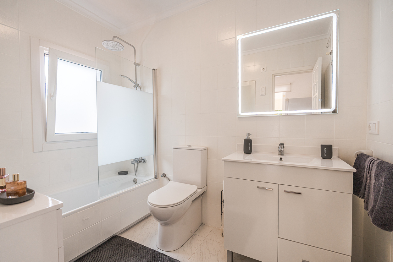Modernes Badezimmer mit weißen Fliesen, Waschtisch mit LED-hinterleuchtetem Spiegel und Badewanne mit Glasduschwand und Regenduschkopf.