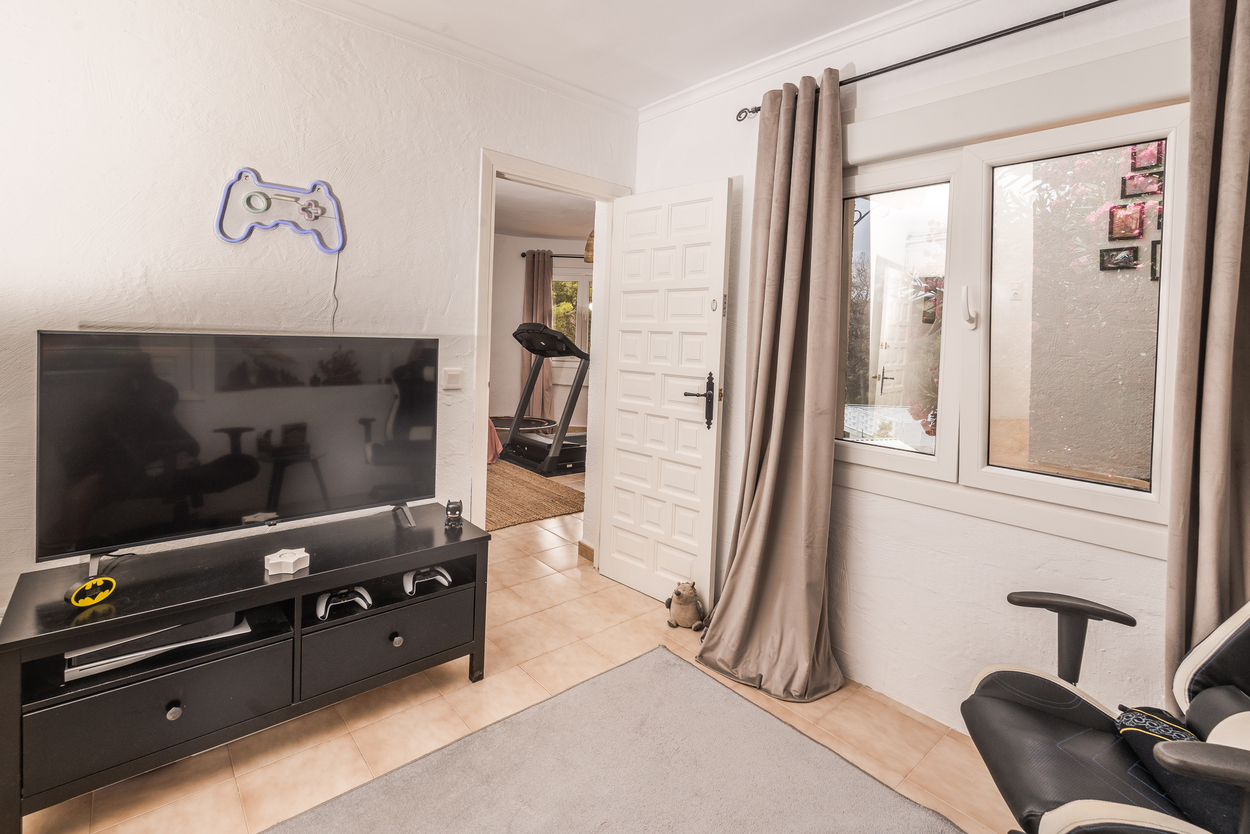 Multimediaraum mit Gaming-Ecke, Flachbildfernseher und Fliesenboden, mit Blick in einen angrenzenden Fitnessbereich inklusive Laufband.
