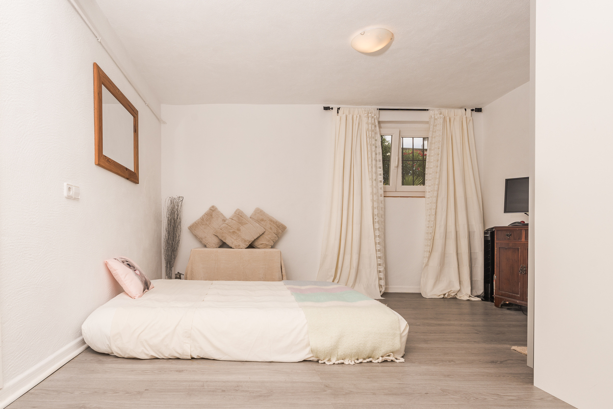 Schlafzimmer mit grauem Laminatboden in Holzoptik, weißen Wänden und niedrigem Bett. Mit dunklem Holzschrank, Holzspiegel und Fenster mit hellen Vorhängen.