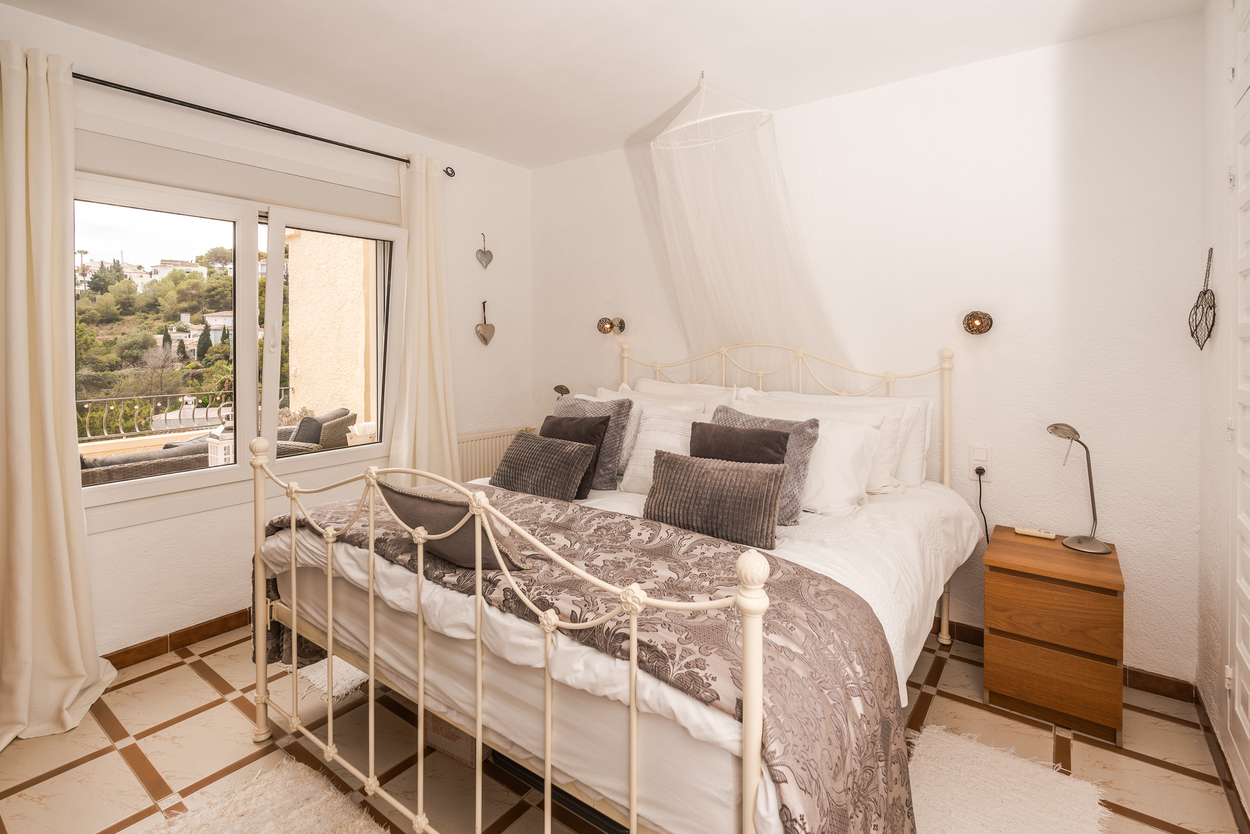 Schlafzimmer mit weißem Metallbett, dekorativem Fliesenboden und doppelt verglastem Fenster. Der Raum bietet Ausblick auf die Natur und verfügt über einen Heizkörper.