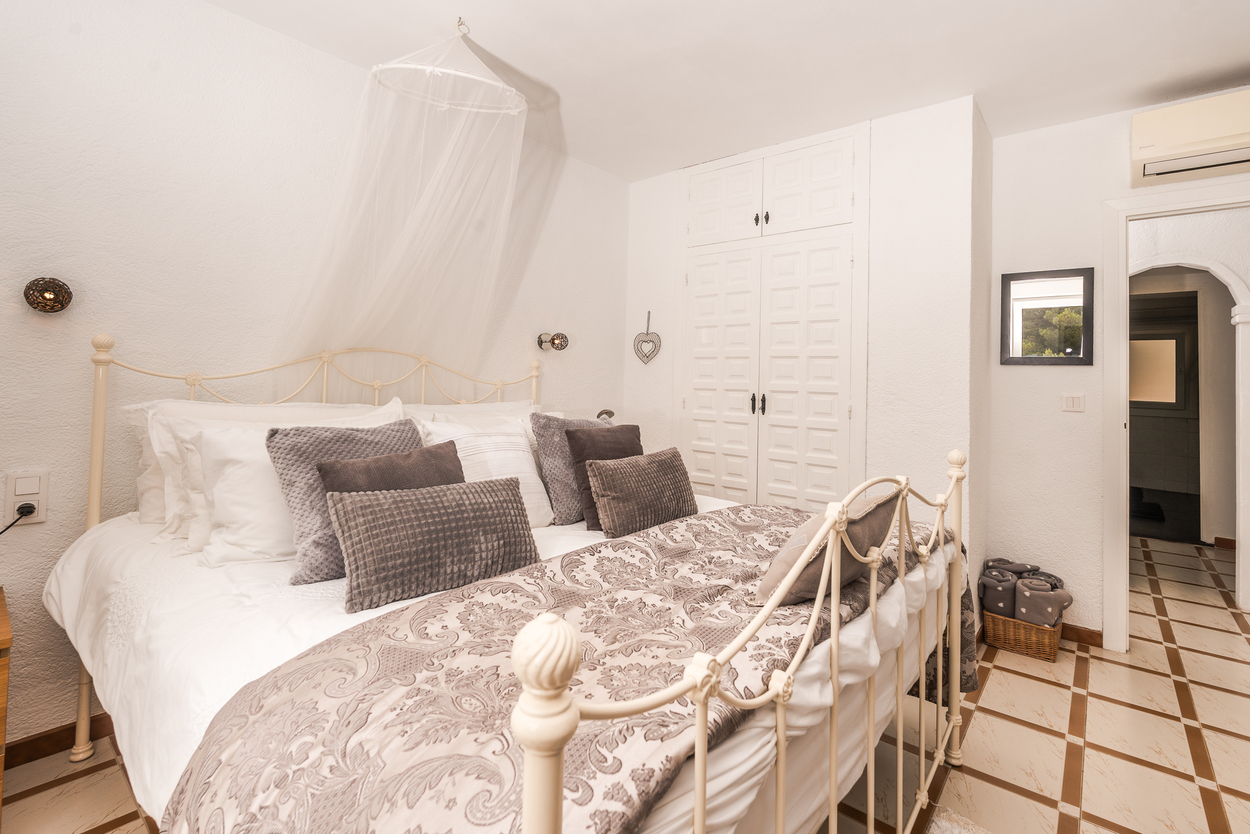 Schlafzimmer mit weißem Schmiedeeisenbett, Moskitonetz und Einbauschränken. Mit Fliesenboden und Klimaanlage über dem bogenförmigen Durchgang.