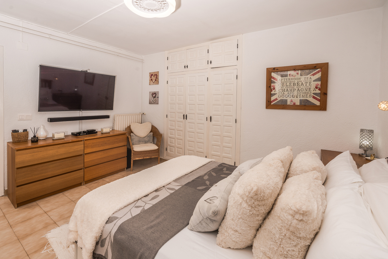 Schlafzimmer mit weißen Einbauschränken, großem Wand-TV über einer Holzkommode und Fliesenboden. Ausgestattet mit Heizkörper und einem Korbsessel.