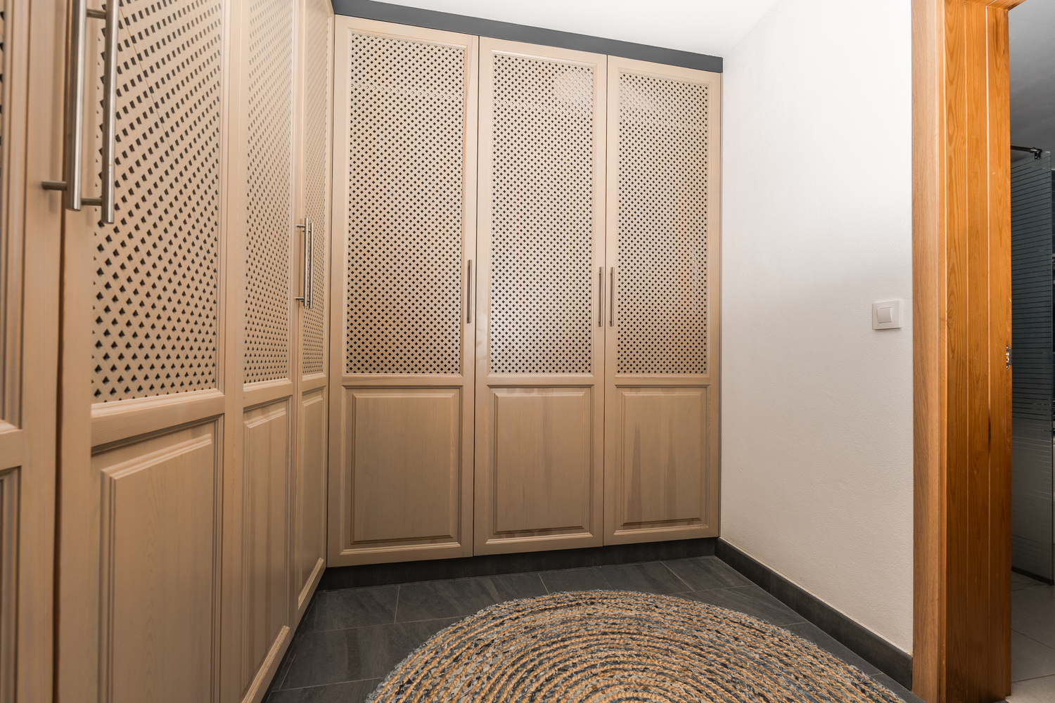 Ankleidezimmer mit Einbauschränken aus hellem Holz und Gitterpaneelen, dunklem Fliesenboden und einem runden Jute-Teppich.
