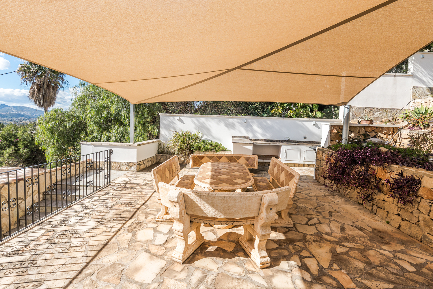 Außen-Essbereich mit kunstvoll gefertigtem Steintisch und Bänken, beschattet durch ein beiges Sonnensegel auf einer Naturstein-Terrasse neben der Sommerküche.