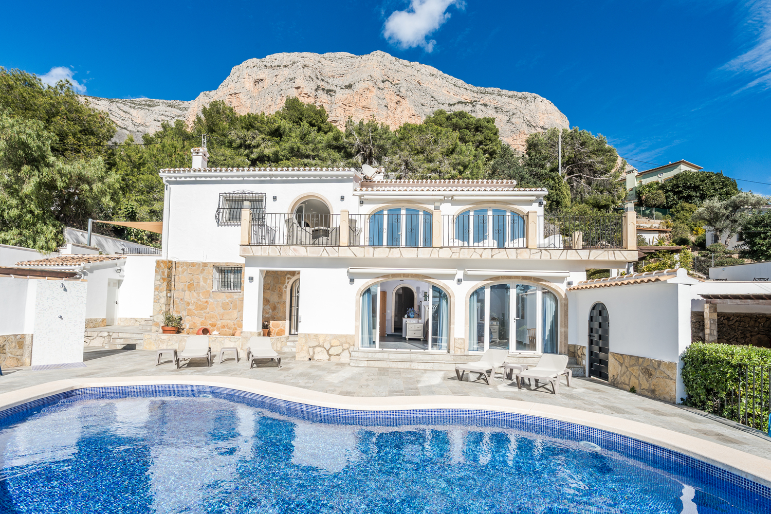 Außenansicht der Villa mit geschwungenem Pool, Natursteinterrasse und Rundbogenfenstern direkt unter dem markanten Felsmassiv des Montgó.