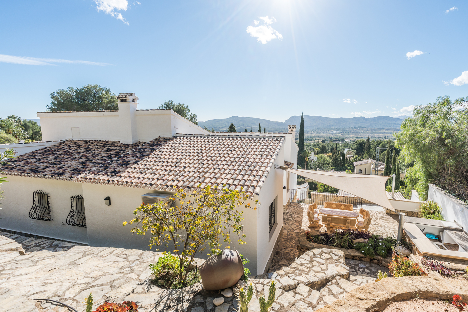Außenansicht der Villa mit traditionellem Ziegeldach, Natursteinterrassen, einem Essbereich unter einem Sonnensegel und weitem Panoramablick auf die Berge.