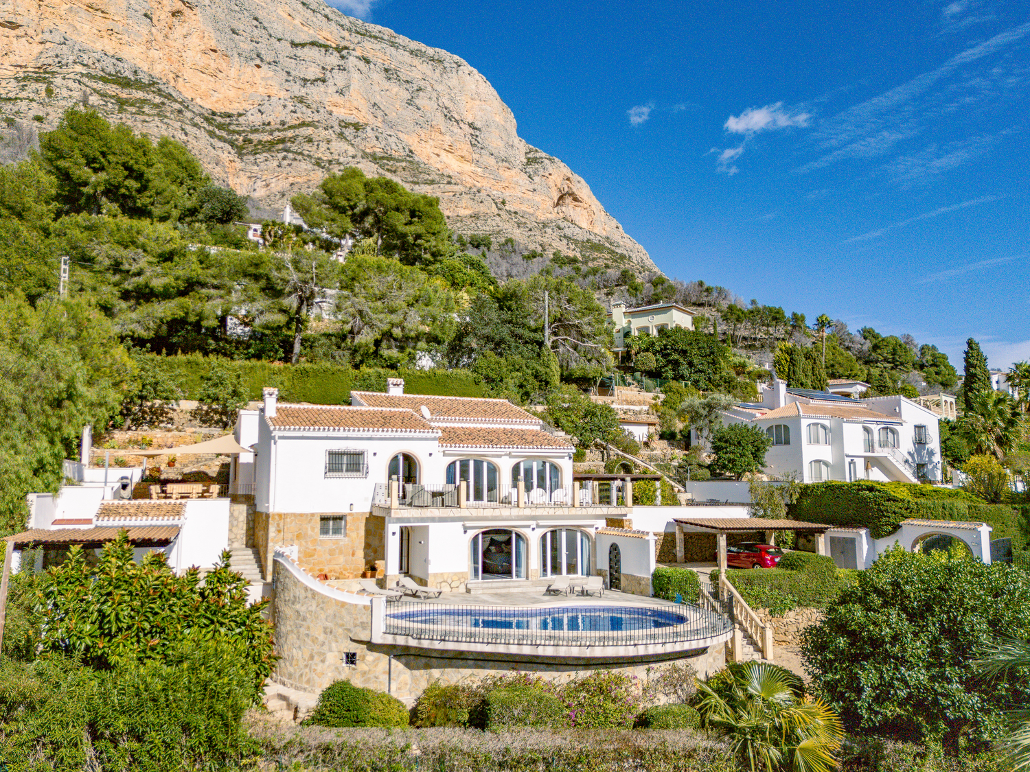 Außenansicht der zweistöckigen Villa am Fuße des Montgó mit geschwungenem Pool, weißen Fassaden mit Natursteinsockel und Rundbogenfenstern auf den Terrassen.
