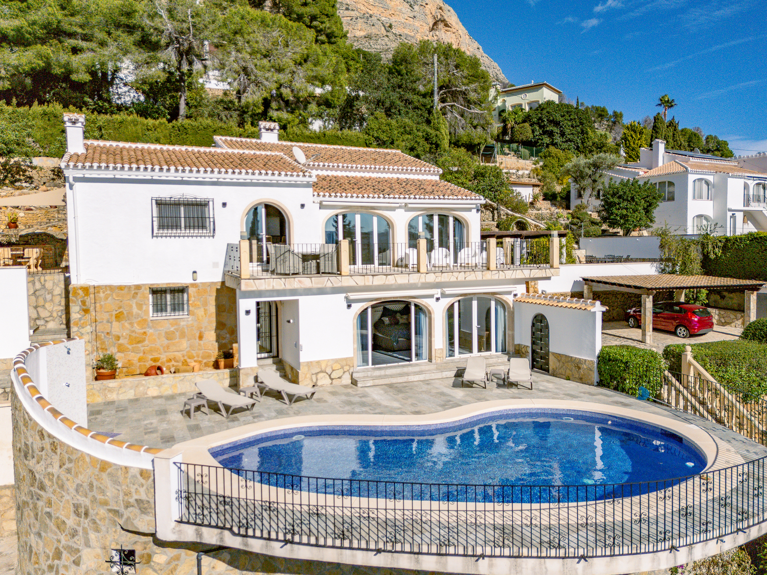 Außenansicht der zweistöckigen Villa mit nierenförmigem Pool, Natursteinterrasse und Rundbogenfenstern vor der Kulisse des Montgó-Massivs.