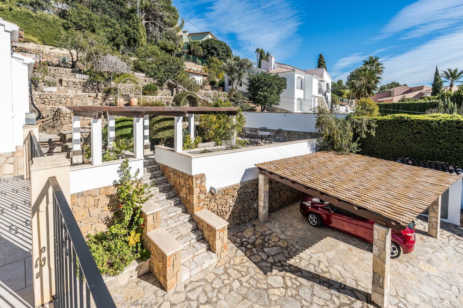 Außenansicht mit gepflasterter Einfahrt, Carport mit Ziegeldach und terrassiertem mediterranem Garten mit Natursteinmauern und Treppen.