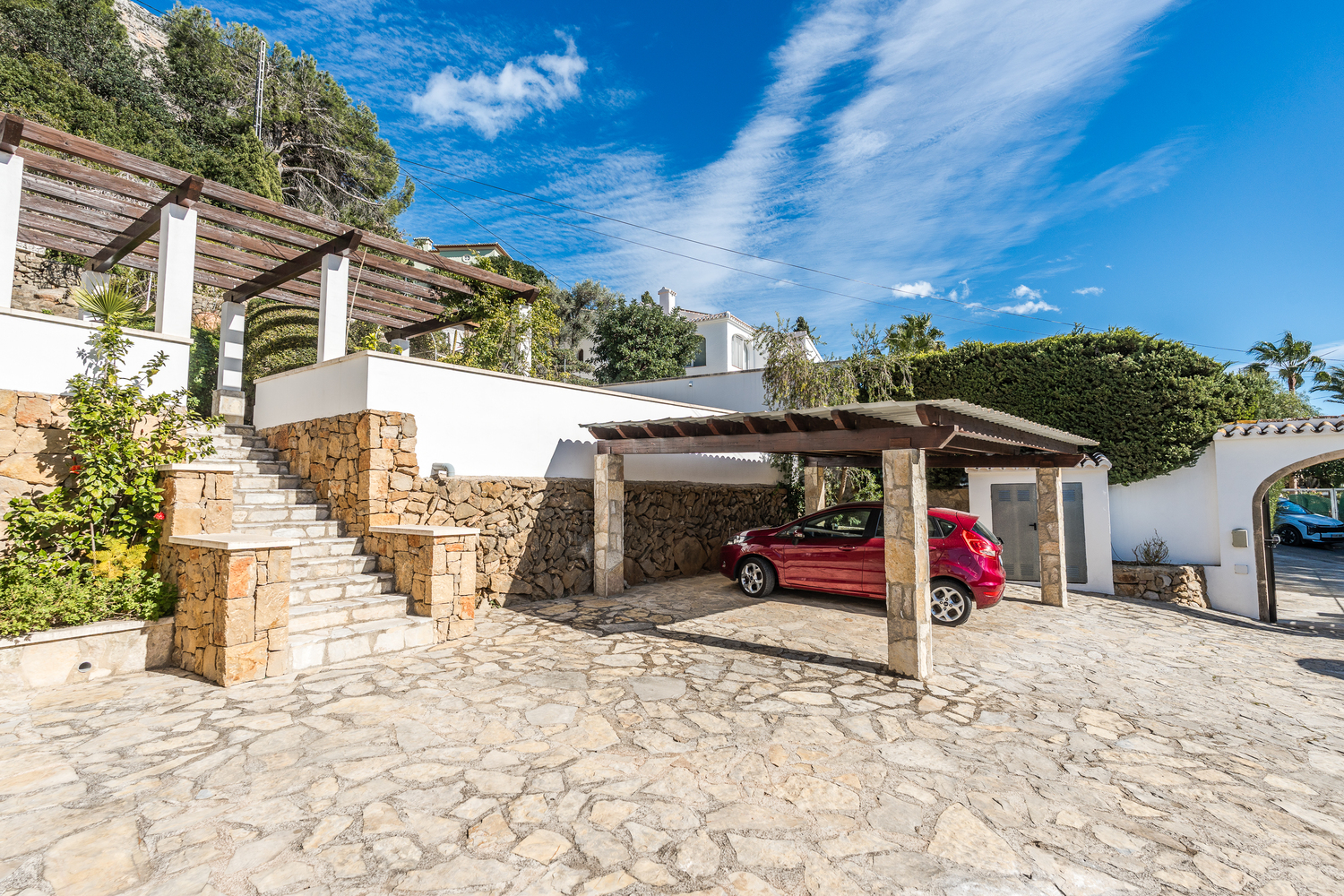 Außenbereich mit Natursteinpflaster, überdachtem Carport und Steintreppen, die zu den verschiedenen Ebenen und Pergolen der Villa führen.