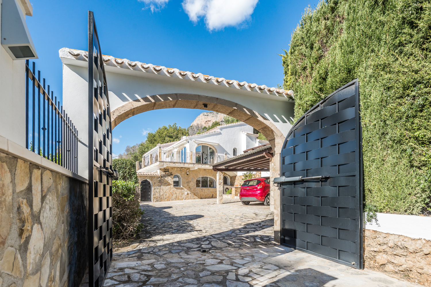 Eingang mit automatischen schwarzen Metalltoren und traditionellem Tosca-Steinbogen, der auf eine gepflasterte Einfahrt mit Blick auf die Villa und den Carport führt.