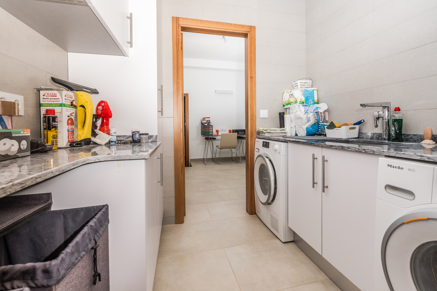 Funktionaler Hauswirtschaftsraum mit weißen Schränken, grauen Marmorarbeitsplatten und Miele-Geräten, mit Blick auf das angrenzende Arbeitszimmer.