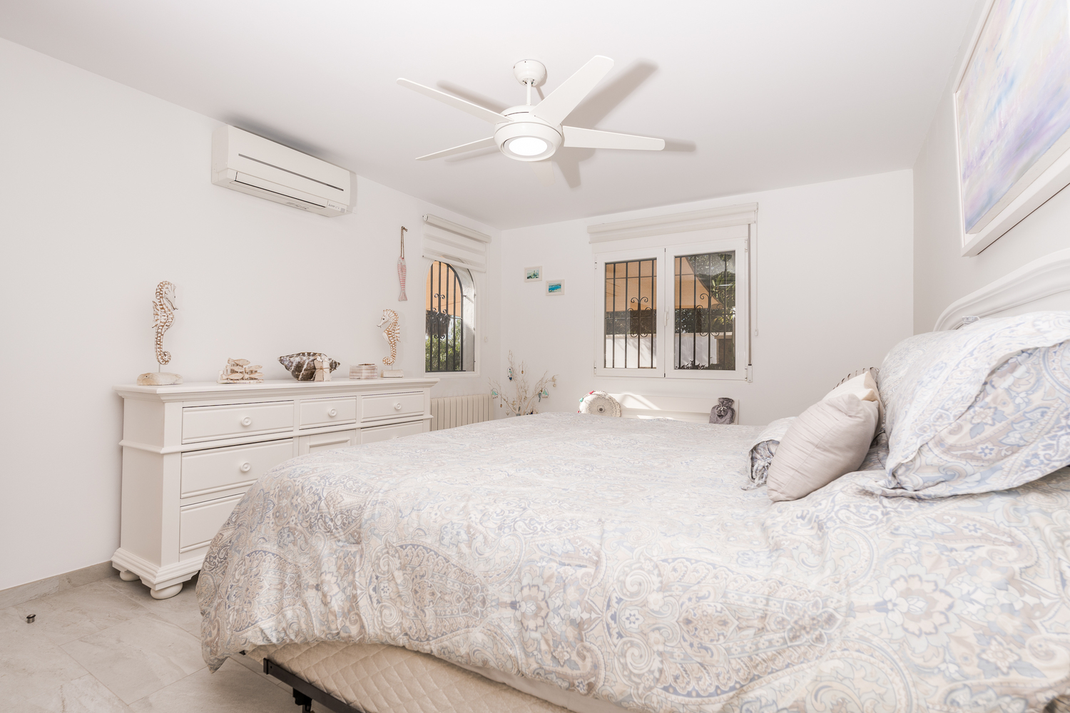 Helles Schlafzimmer mit weißen Wänden, Deckenventilator, Klimaanlage und weißer Kommode. Fenster mit Sicherheitsgittern und Fliesenboden ergänzen den Raum.