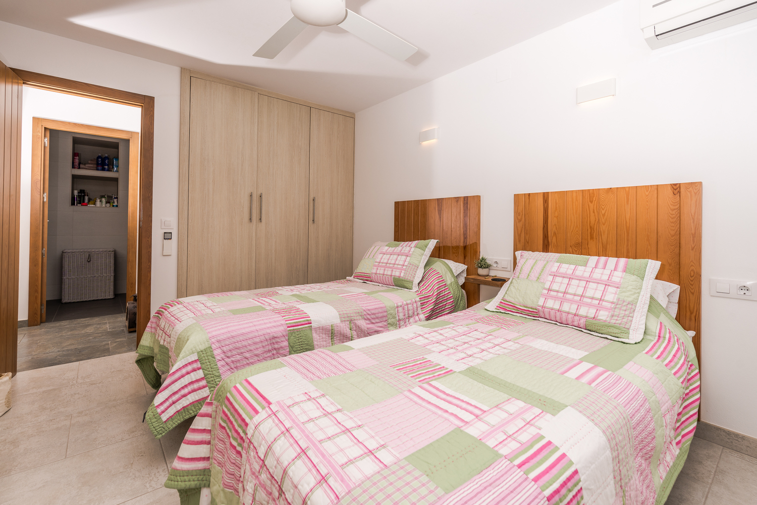 Helles Schlafzimmer mit zwei Einzelbetten, Kopfteilen aus Naturholz, Einbauschränken, Deckenventilator und Zugang zum angrenzenden Badezimmer.