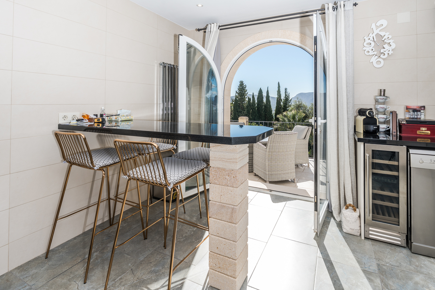 Moderne Frühstücksbar mit schwarzer Granitplatte und goldenen Hockern, die durch einen Rundbogen auf eine Terrasse mit Blick auf das Tal und die Berge führt.