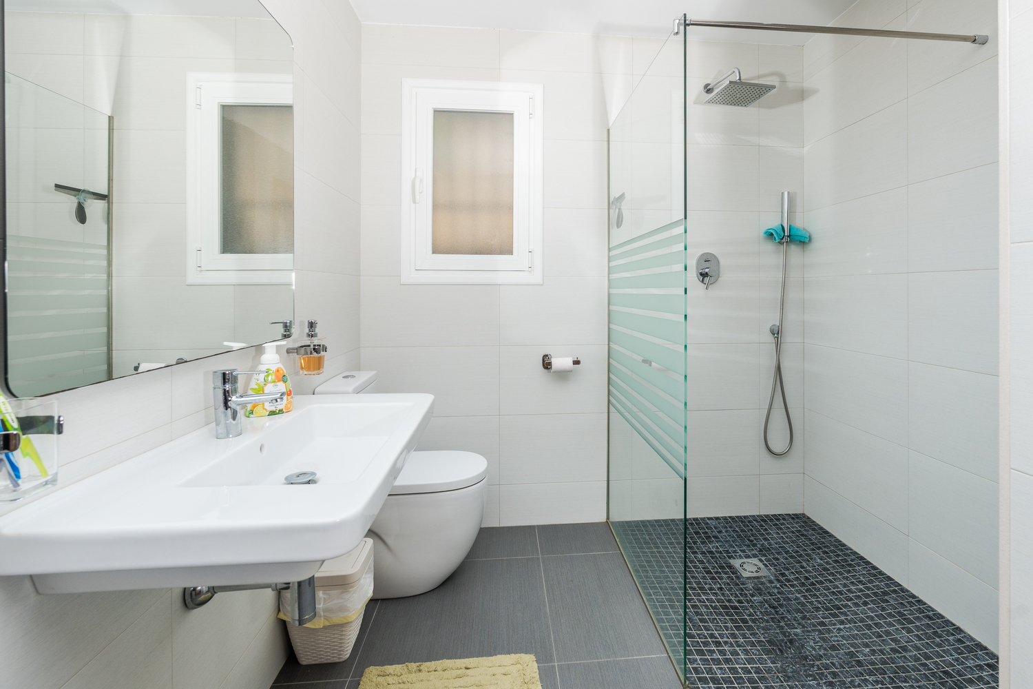 Modernes Badezimmer mit begehbarer Dusche, Glastrennwand und dunklem Mosaikboden. Ausgestattet mit einem Wandwaschbecken und hellen Fliesen für ein helles Ambiente.
