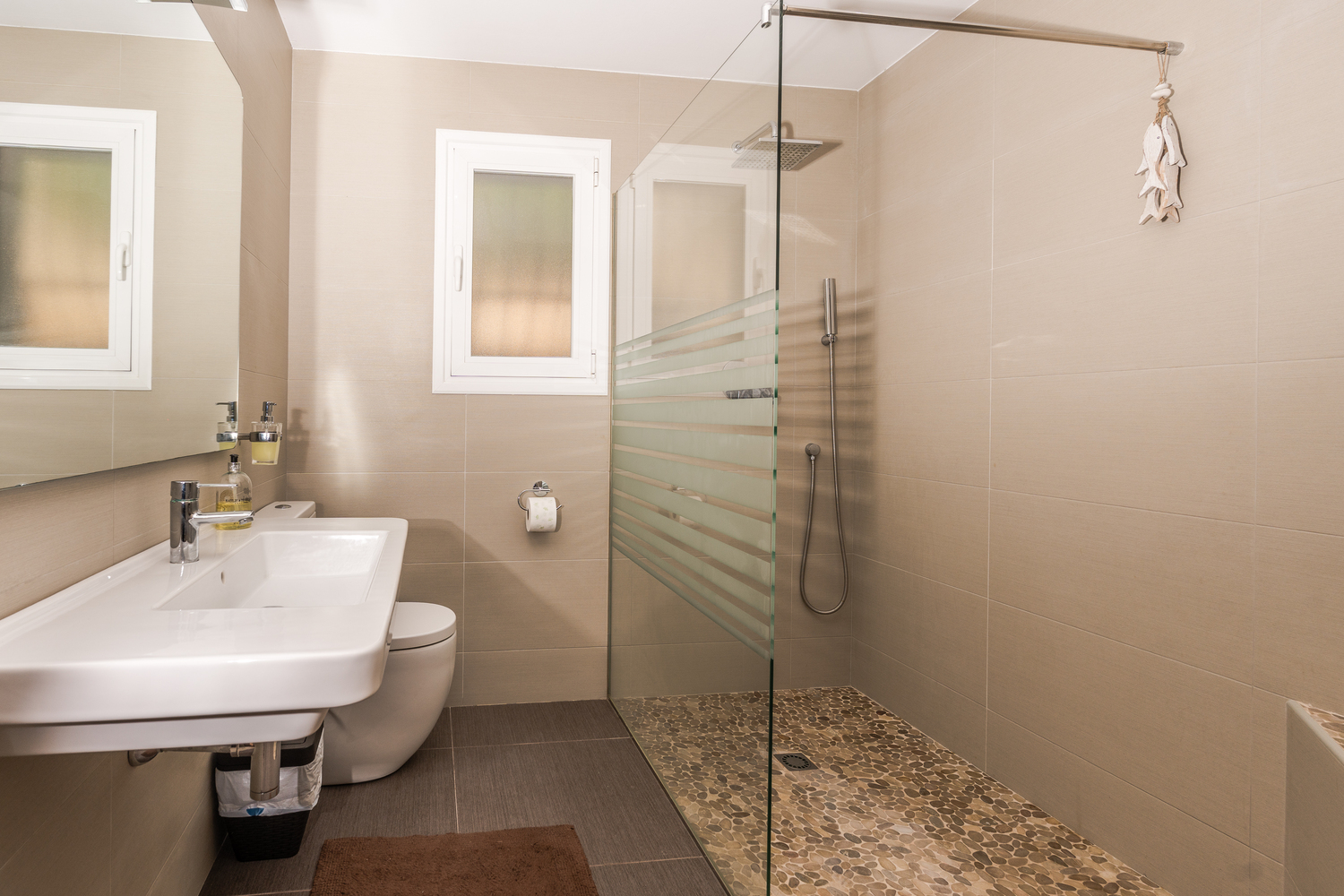 Modernes Badezimmer mit begehbarer Dusche, Kieselsteinboden, Glastrennwand und neutralen Fliesen. Ausgestattet mit einem Wandwaschbecken und Milchglasfenster.