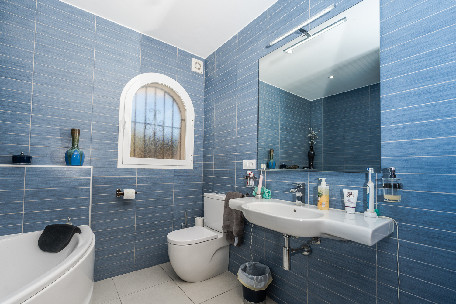 Modernes Badezimmer mit blau strukturierten Wandfliesen, einem Rundbogenfenster mit Sichtschutzglas, weißem Waschbecken und großem Spiegel mit Beleuchtung.
