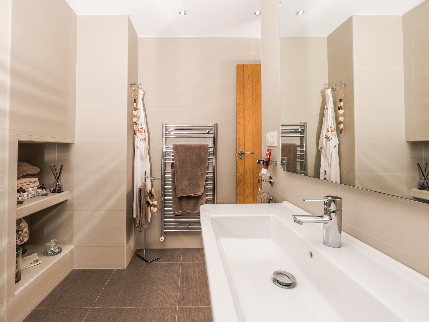 Modernes Badezimmer mit großem Keramikwaschbecken, Wandnischenregalen und verchromtem Handtuchheizkörper vor neutralen beigen Fliesen.