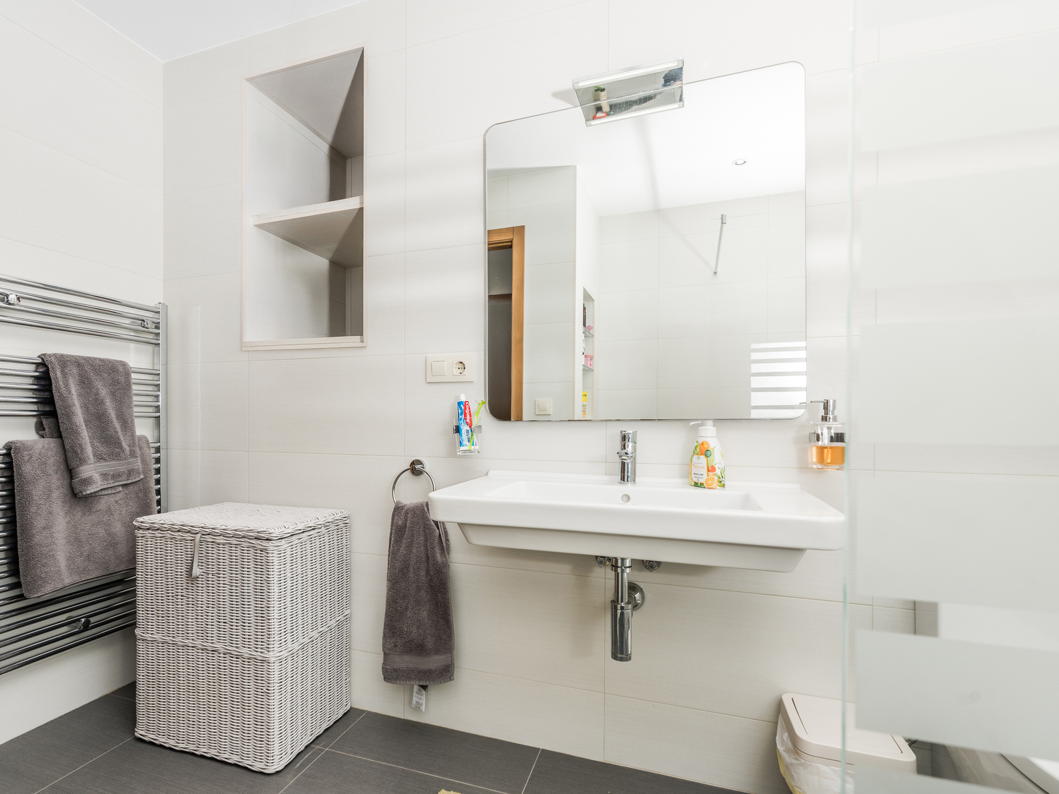 Modernes Badezimmer mit wandhängendem Waschbecken, integrierter Wandnische und verchromtem Handtuchheizkörper auf weißen Fliesen.
