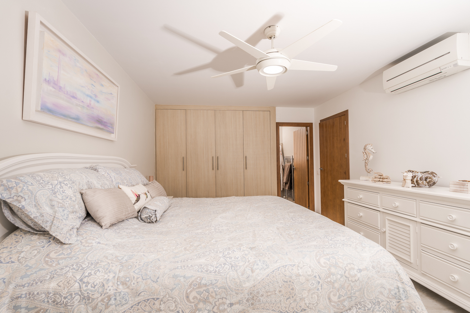 Modernes Schlafzimmer mit Einbauschränken aus hellem Holz, weißer Kommode und Deckenventilator. Ausgestattet mit Klimaanlage und Zugang zum En-Suite-Bereich.
