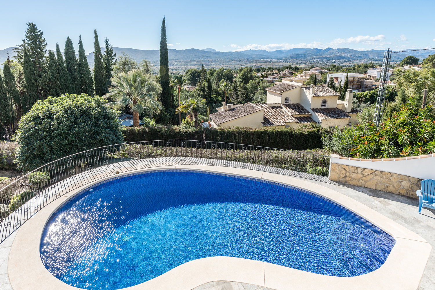 Nierenförmiger Swimmingpool mit blauen Mosaikfliesen, umgeben von einer Steinterrasse und einem Schmiedeeisengeländer, mit Panoramablick auf das Tal und die Berge.