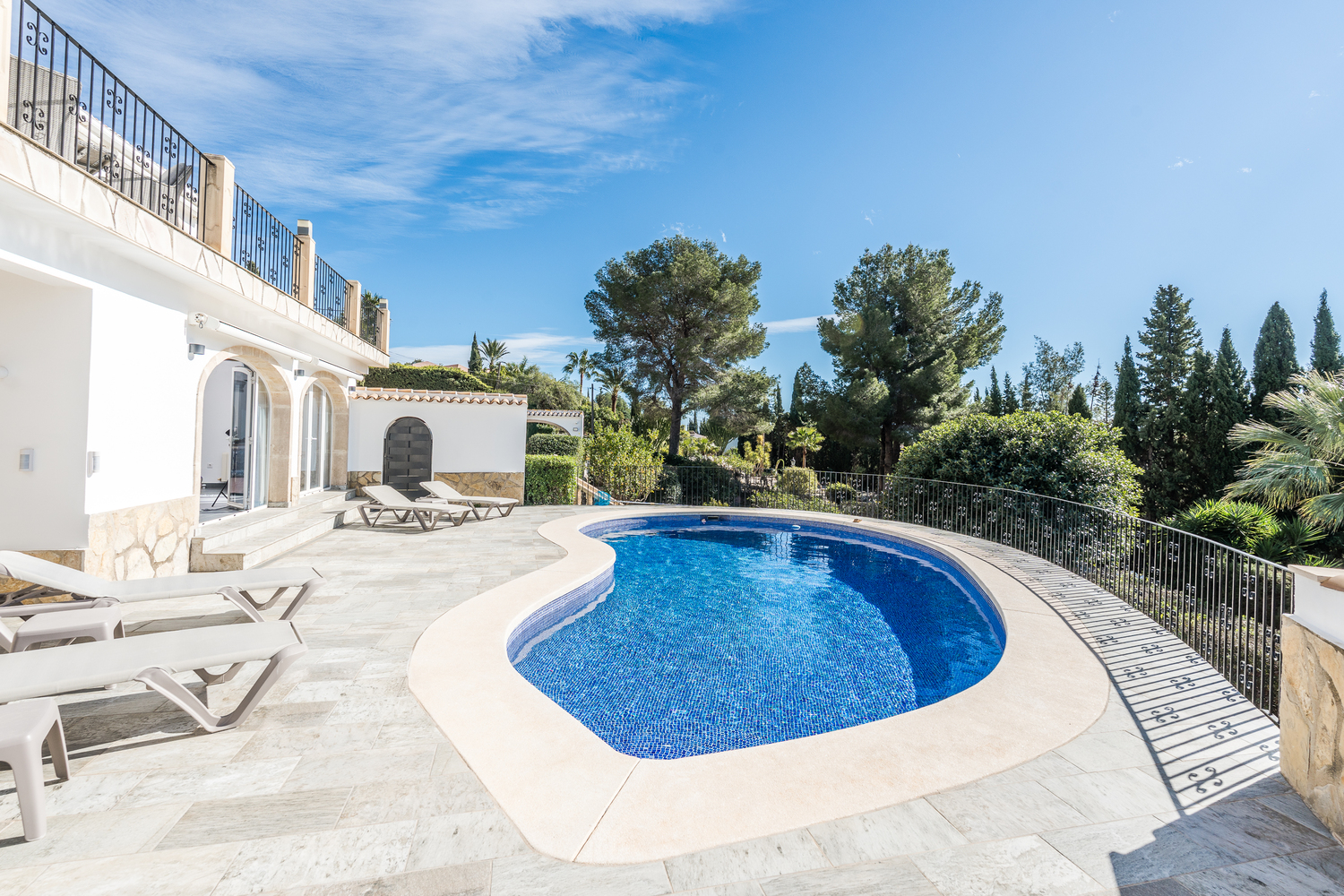 Nierenförmiger Swimmingpool, umgeben von einer hellen Steinterrasse mit Sonnenliegen, eingebettet in mediterrane Vegetation vor der weißen Villenfassade.