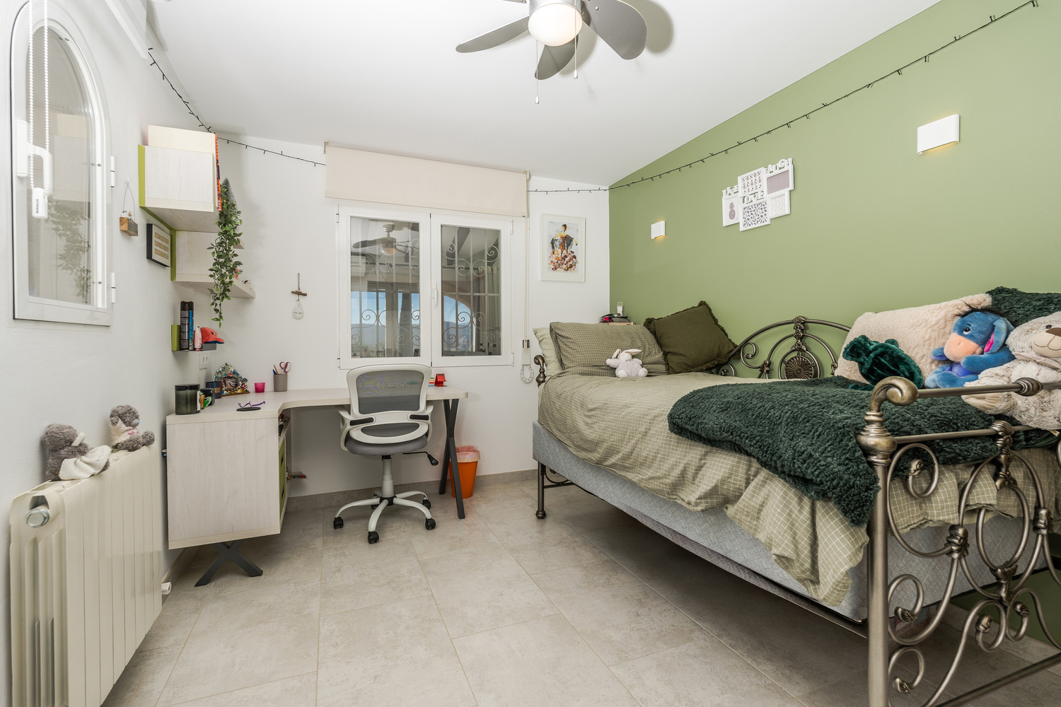 Schlafzimmer mit grüner Akzentwand, ornamentalem Metallbettrahmen und integriertem Arbeitsbereich mit Schreibtisch. Ausgestattet mit Deckenventilator und Heizkörper.